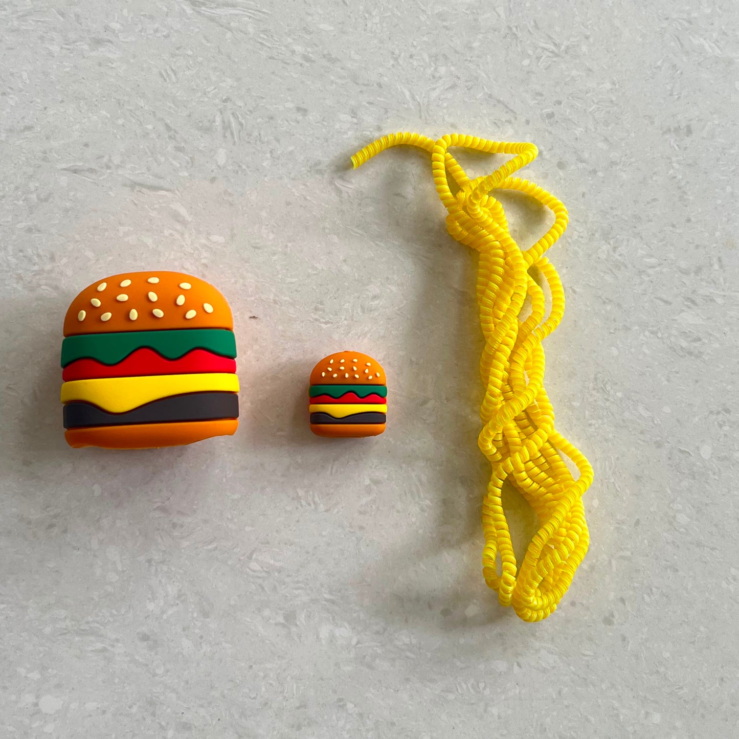 A pair of miniature plastic earbuds case covers shaped like burgers, shown next to a bright yellow braided cord on a light gray textured background.