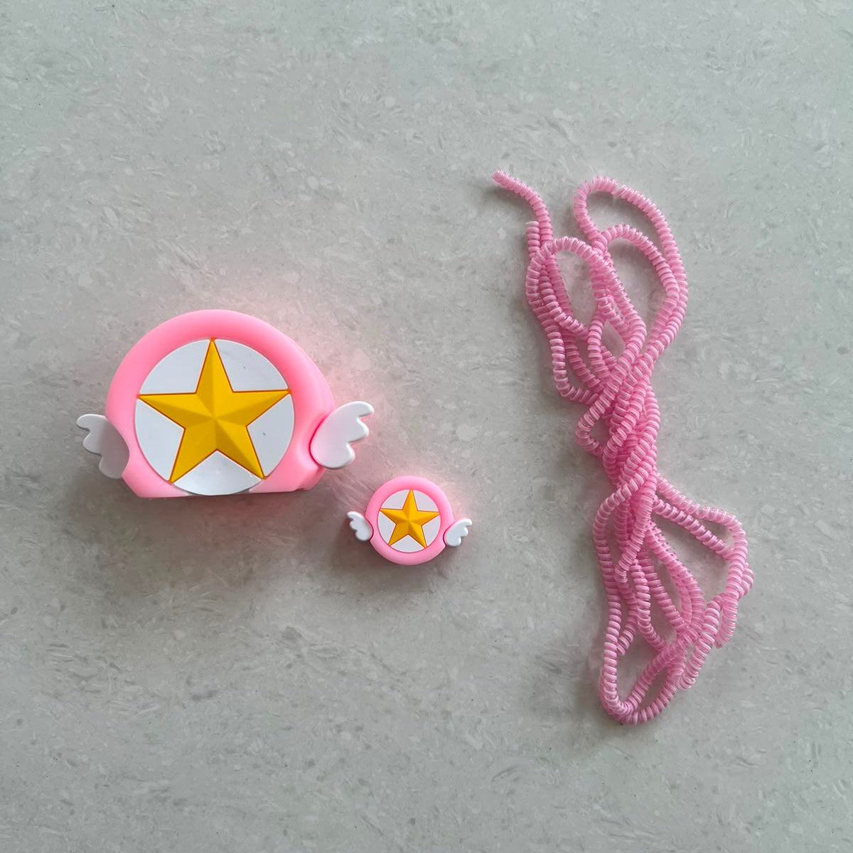 Pink winged star charger adapter cover, mini cable protector and pink spiral cable wrap components flat lay on grey surface