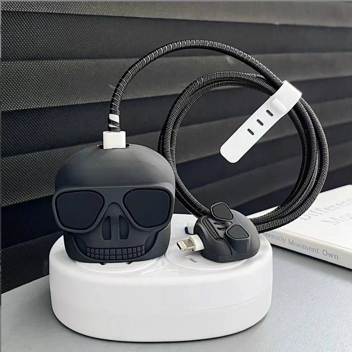 A black skull-shaped USB charger on a white charging dock, with a coiled black cable and a white plug adapter, shown on a gray surface against a dark textured background.