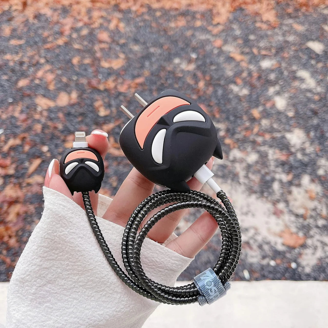 Dark masked hero charger adapter cover and mini cable protector held in hand showing back detail with autumn leaves background