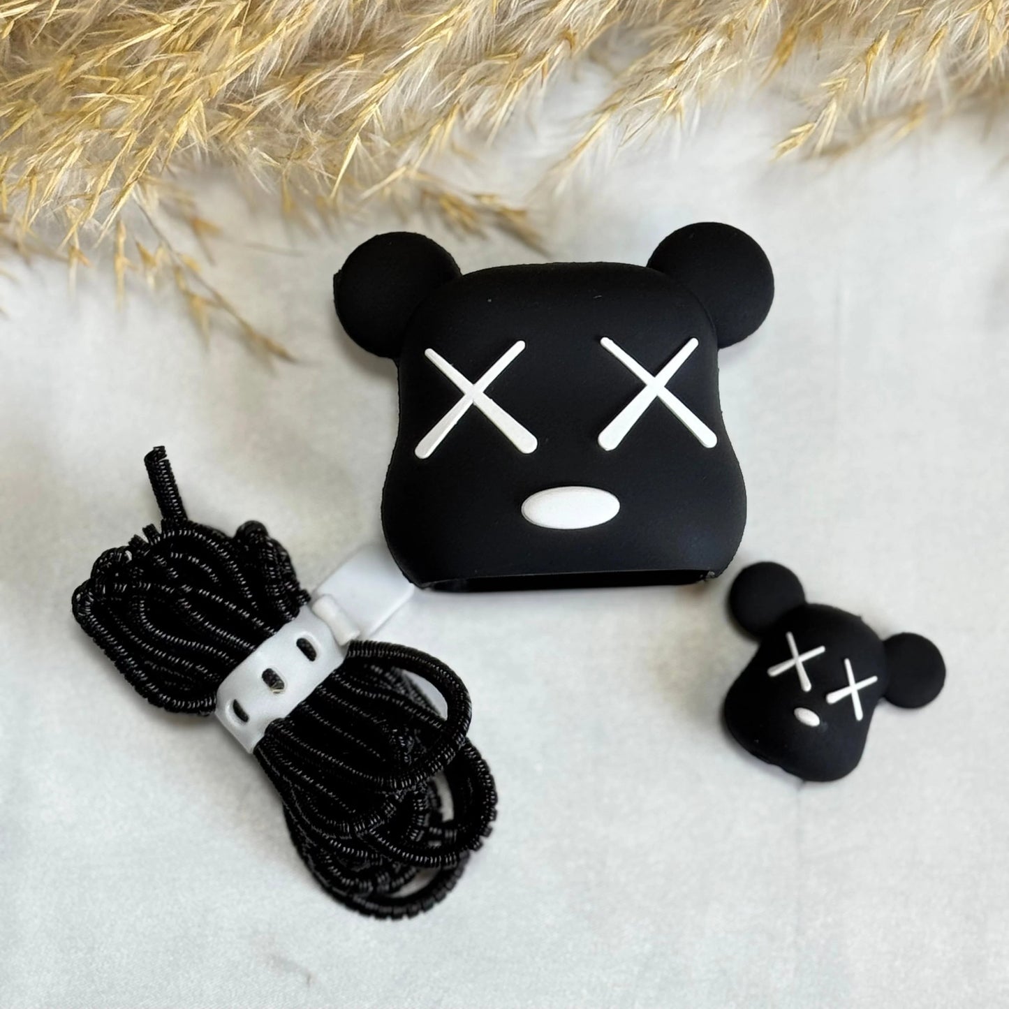 Black silicone AirPods case cover in aKA Bear face design with white embroidered eyes and mouth, shown with a coiled black cable and a small matching black accessory on a white surface with dried pampas grass.