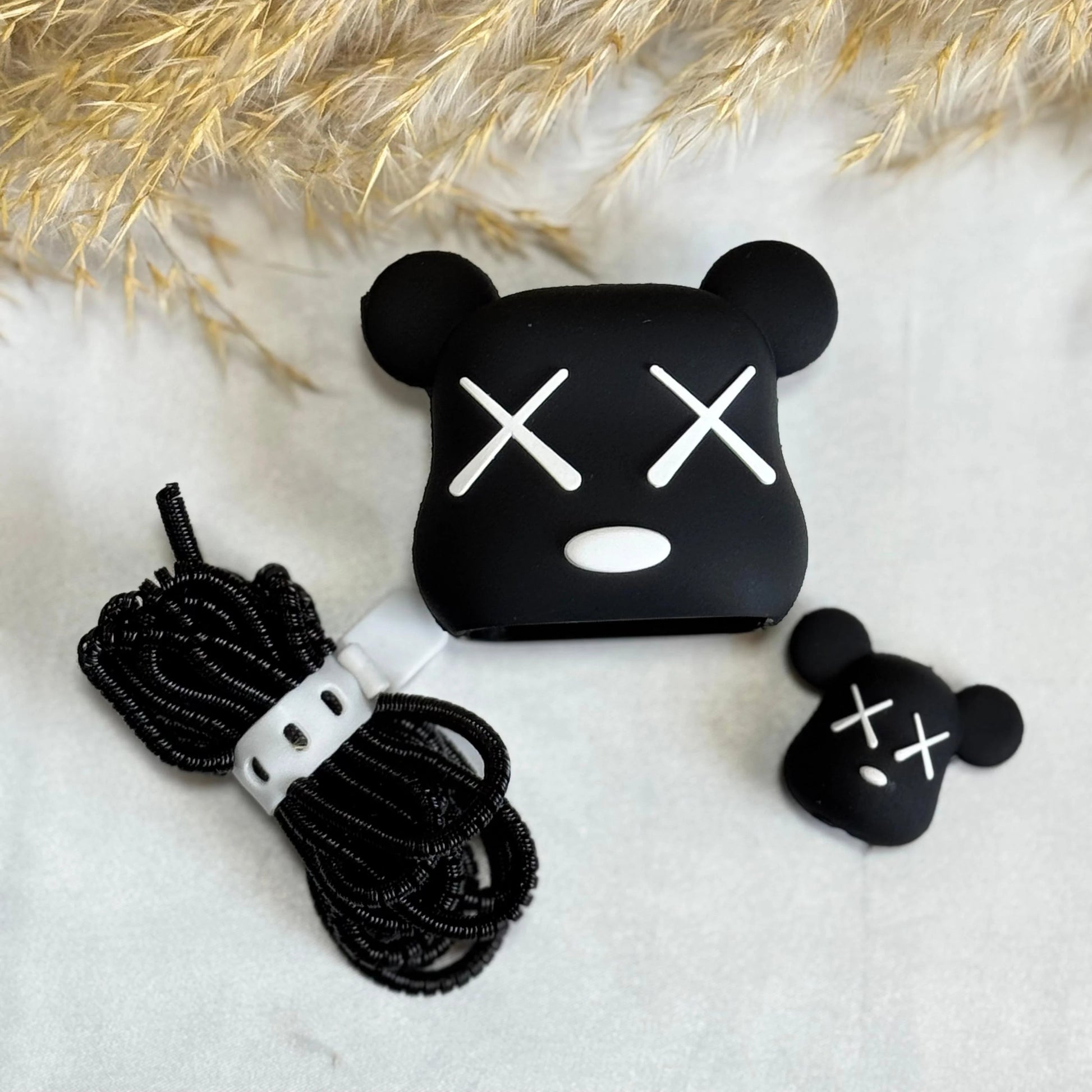 Black silicone AirPods case cover in aKA Bear face design with white embroidered eyes and mouth, shown with a coiled black cable and a small matching black accessory on a white surface with dried pampas grass.