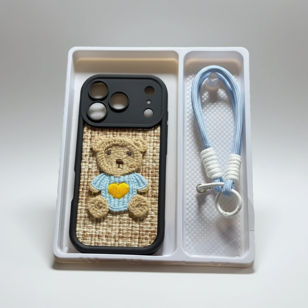 A hand holds a white plastic tray containing a black phone case with a woven tan panel and a teddy bear applique, alongside a light blue braided wrist strap with white beads and a silver clasp. The background is a softly blurred indoor setting.