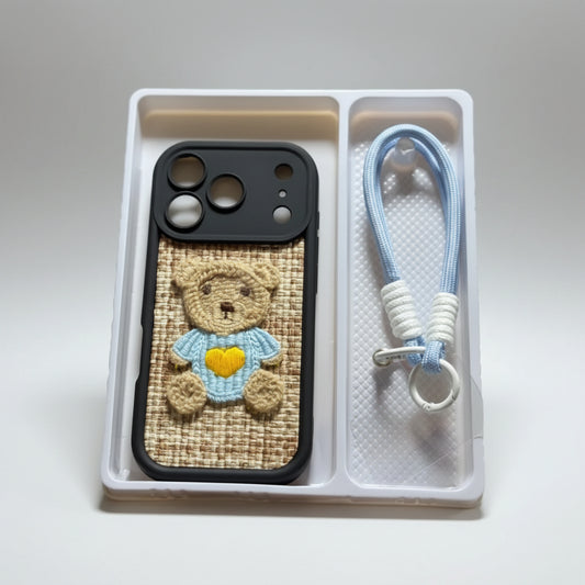 A hand holds a white plastic tray containing a black phone case with a woven tan panel and a teddy bear applique, alongside a light blue braided wrist strap with white beads and a silver clasp. The background is a softly blurred indoor setting.