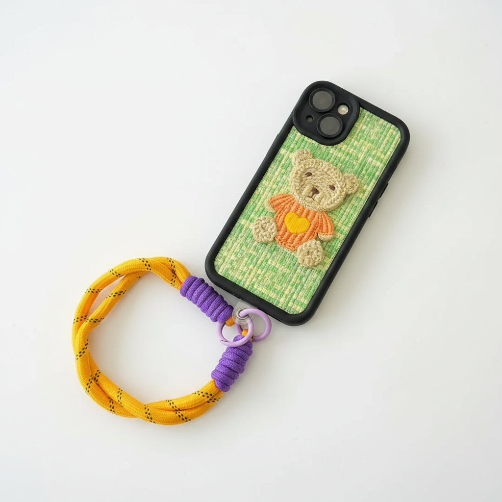 A phone case with a green and yellow checkered background, featuring a hand-embroidered teddy bear wearing an orange shirt. The case is attached to a yellow woven strap with purple accents, laid flat on a black faux fur background.