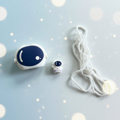 A small white and navy blue astronaut-themed accessory set on a light gray textured surface, including a larger rounded astronaut-shaped piece, a tiny astronaut figure, and a long white cord.