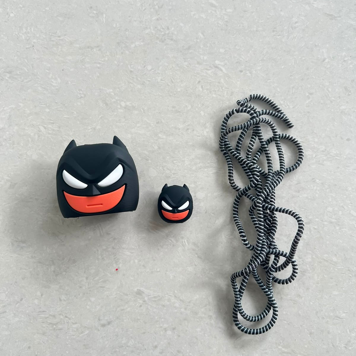 Dark masked hero charger adapter cover, mini cable protector and black spiral cable wrap components flat lay on grey surface