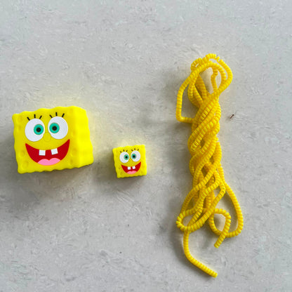 Yellow square smiley character charger adapter cover, mini cable protector and yellow spiral cable wrap components flat lay on grey surface