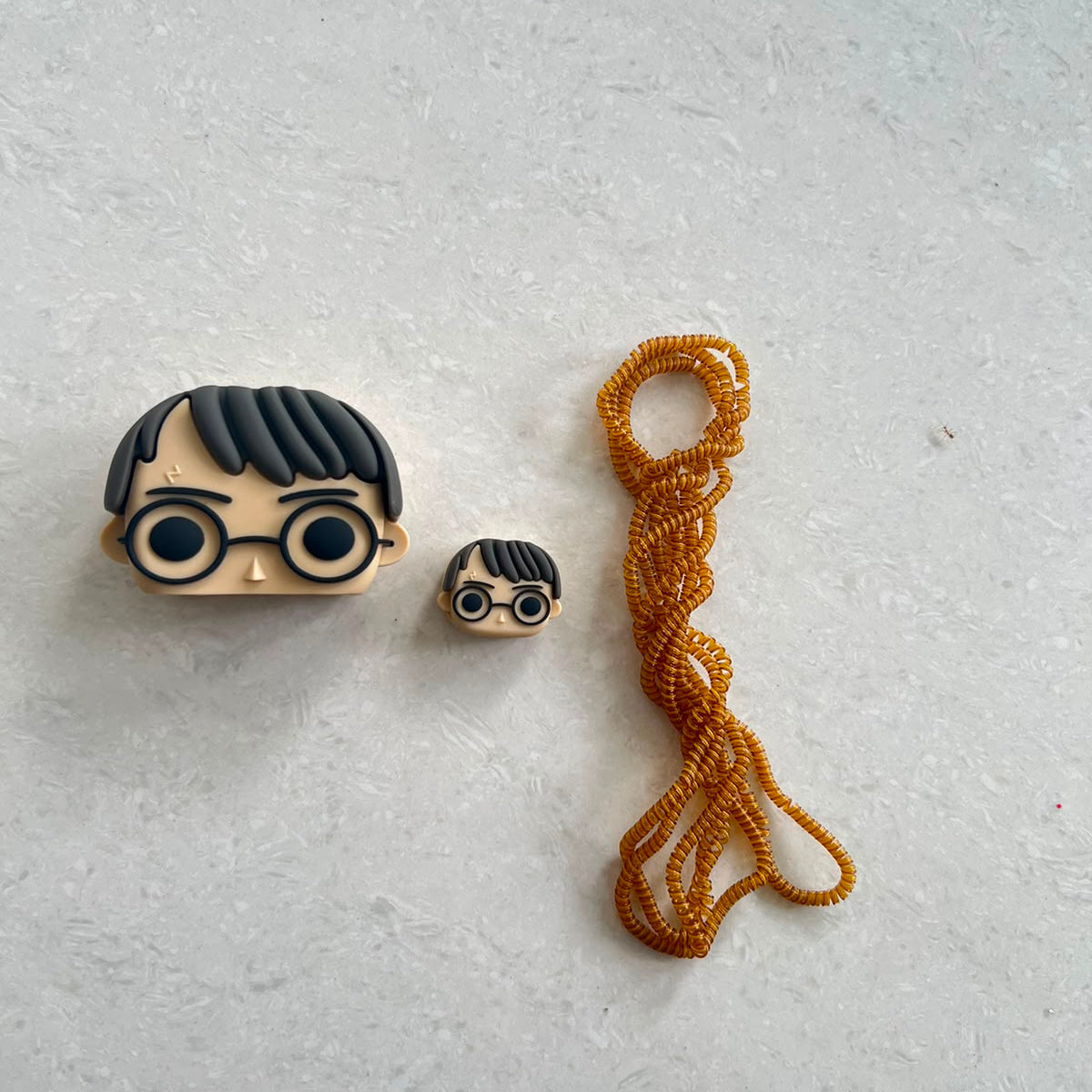 Beige glasses boy wizard charger adapter cover, mini cable protector and gold spiral cable wrap components flat lay on grey surface