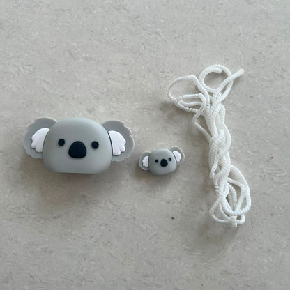Grey koala face charger adapter cover, mini koala cable protector and white spiral cable wrap components flat lay on grey surface