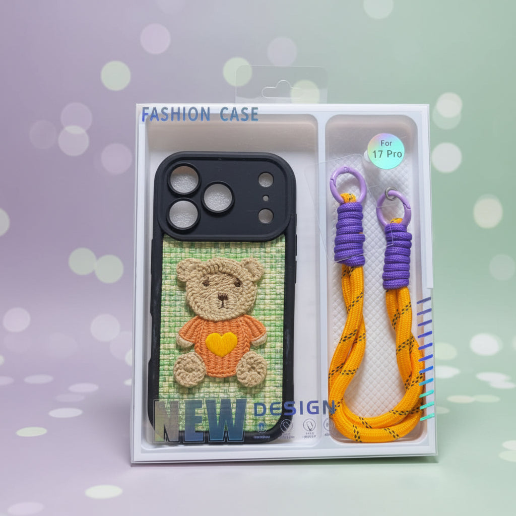 A hand holding a retail package containing a black phone case with a crocheted teddy bear design on a green textured background, alongside two woven wrist straps in purple and orange.